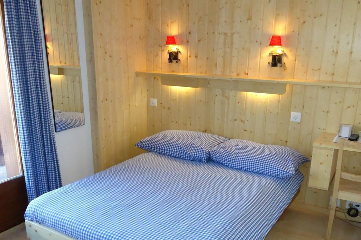 A bedroom with a wooden wall, a bed with checkered bedding, a bedside table, and a mirror.