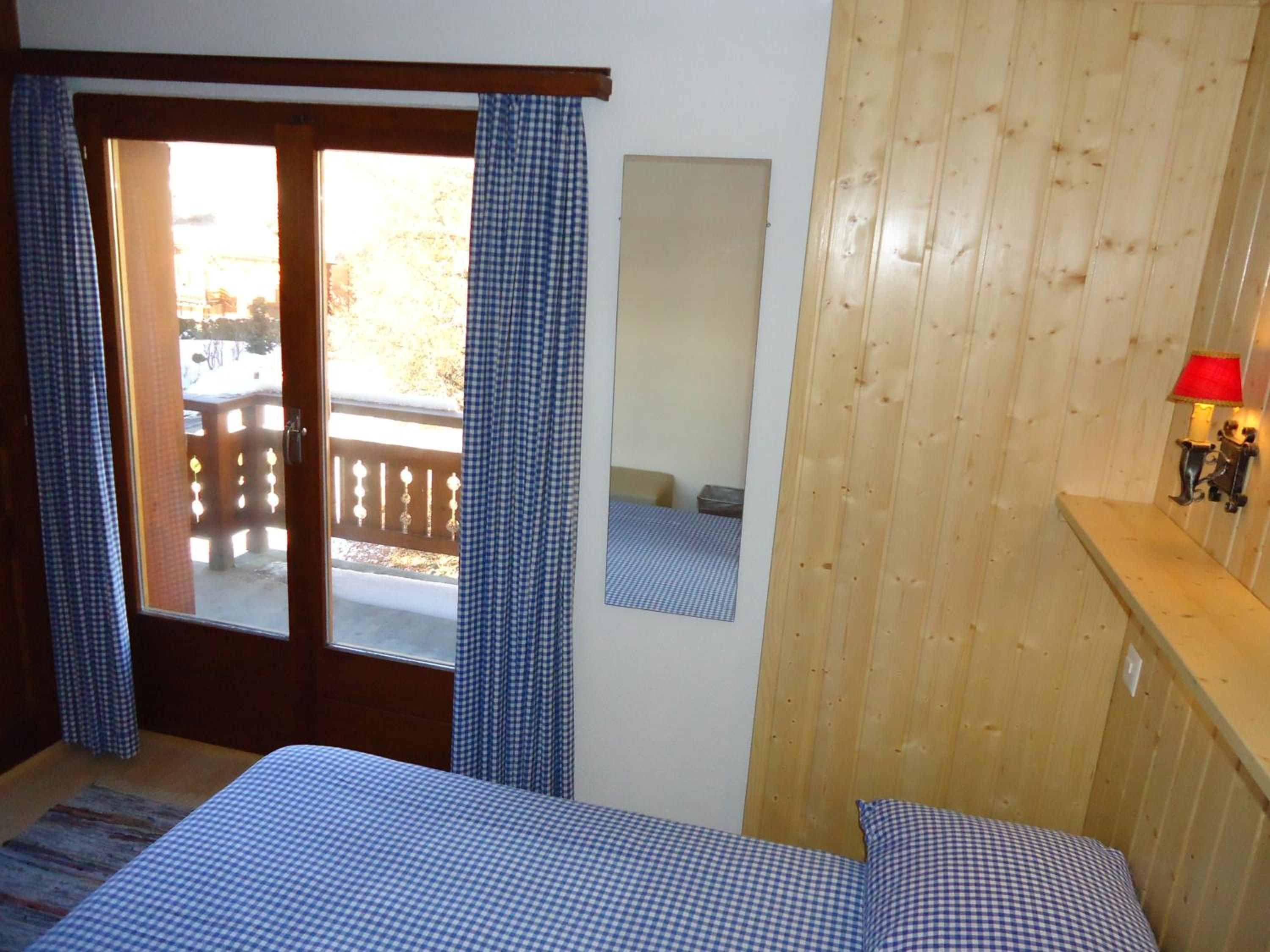 Un dormitorio con pared de madera, una cama con ropa de cama a cuadros, una puerta que da a un espacio exterior y una pequeña lámpara fijada a la pared.