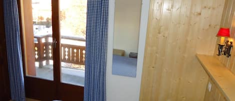 Un dormitorio con pared de madera, una cama con ropa de cama a cuadros, una puerta que da a un espacio exterior y una pequeña lámpara fijada a la pared.
