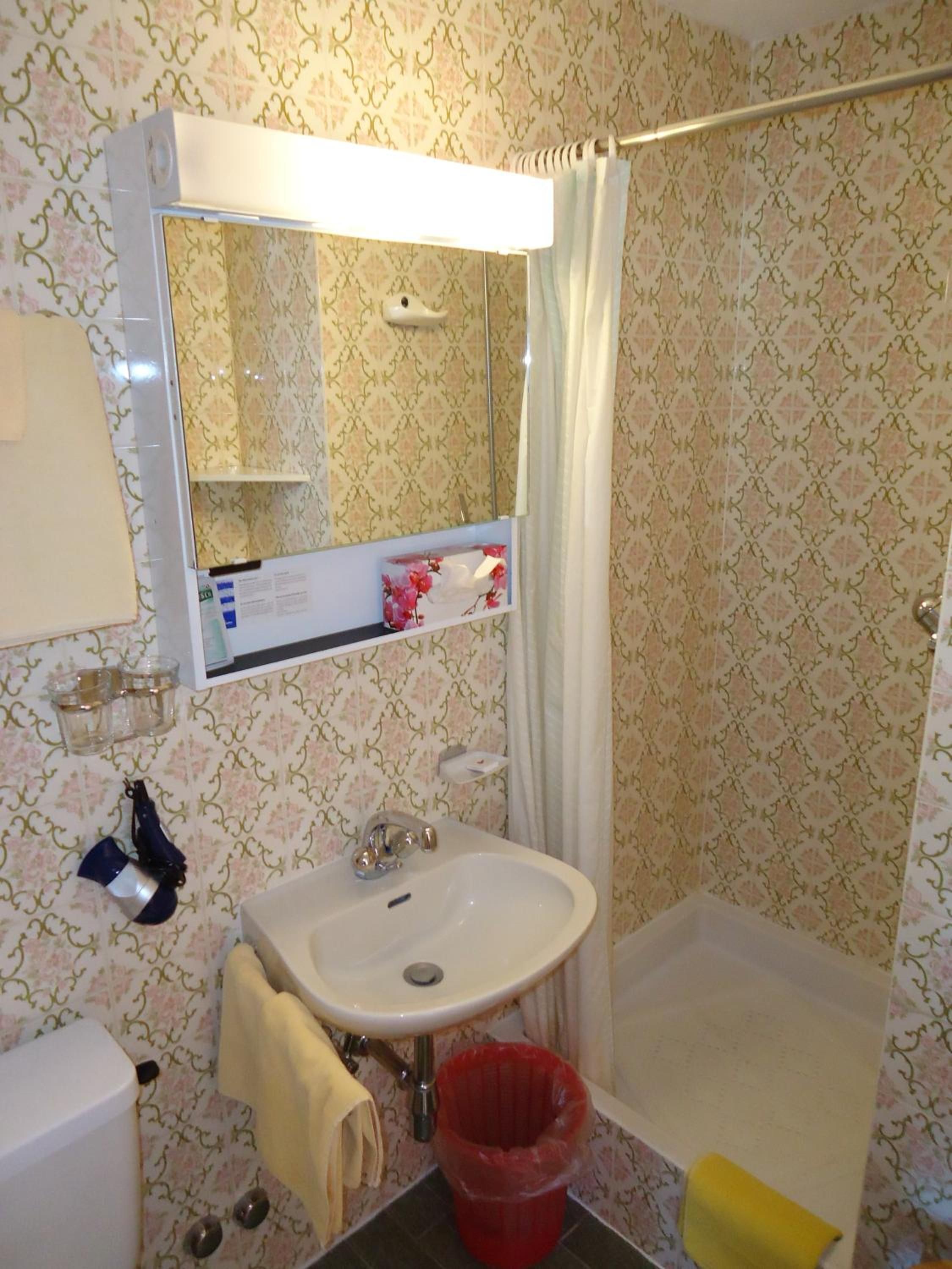 Une salle de bain avec un lavabo blanc, un miroir, un rideau de douche et une poubelle rouge.