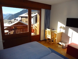 Quarto com cama, escrivaninha, cadeira, televisão e vista para uma montanha.