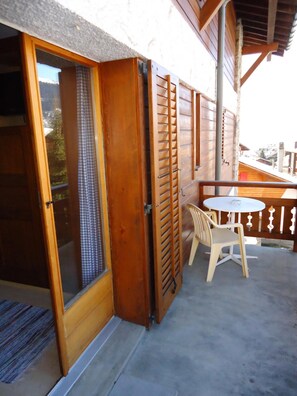 A wooden balcony with a sliding glass door, a small round table, and two plastic chairs. - Hotel Mirabeau (Bagnes)
