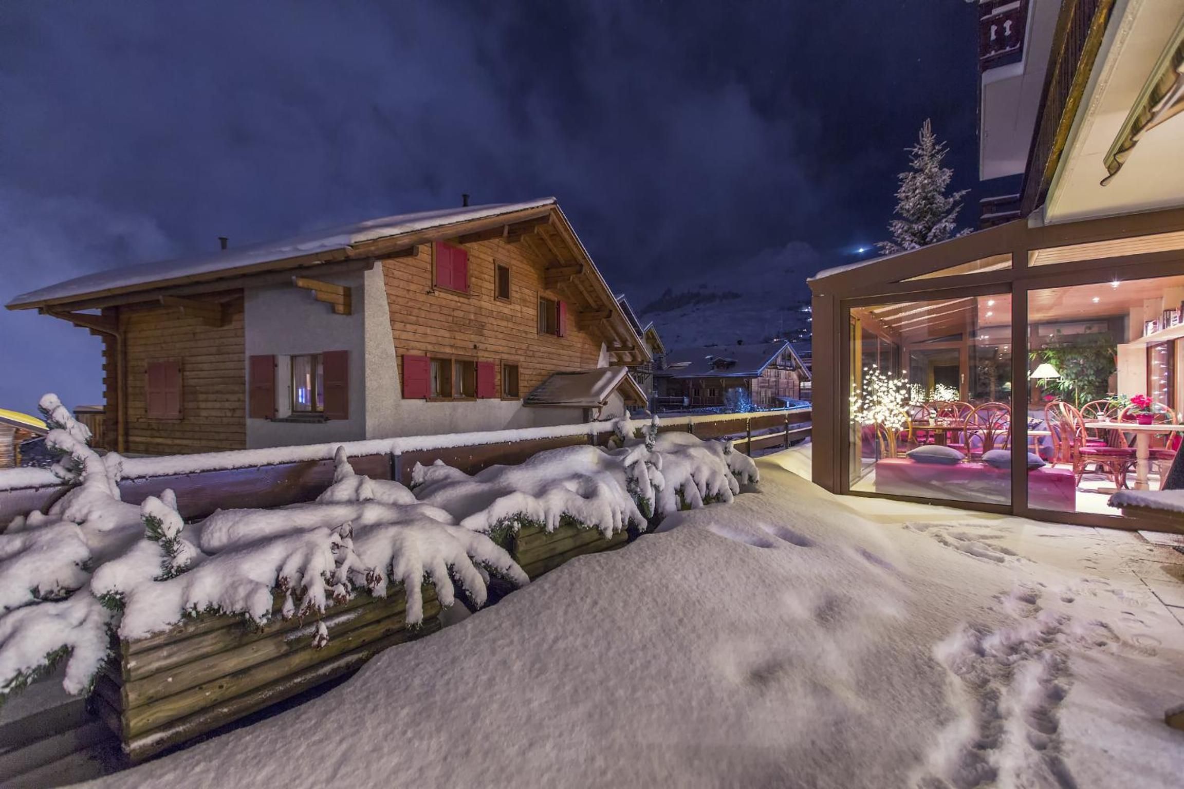 Et snæverkket uteområde med trehus, en glassinnfelt terrasse med møbler og et snødekt landskap.