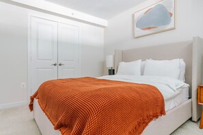 A bedroom with a bed, white walls, a door, a nightstand, and a framed picture on the wall. - Blueground | Ballston, pool, nr Arlington Cemetery (Arlington)