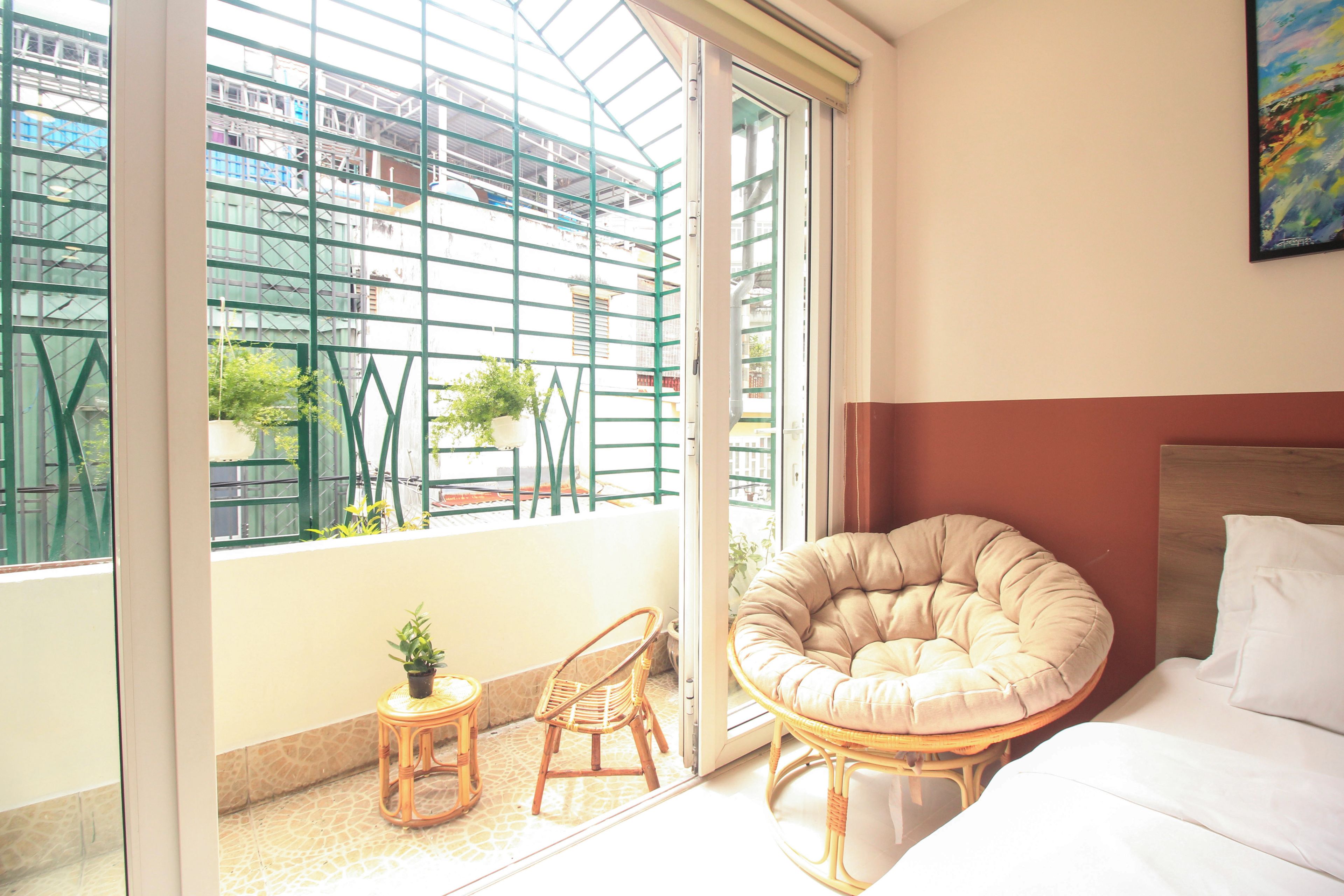 A room with a bed, a round cushioned chair, a small table with a plant, and a view of buildings through a window with a metal grid.