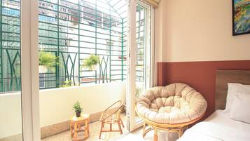 A room with a bed, a round cushioned chair, a small table with a plant, and a view of buildings through a window with a metal grid.