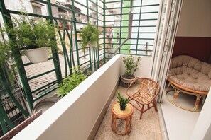 Ein Balkon mit grüner Metallreling, Blumentöpfen, Korbmöbeln und Blick auf die Straße.