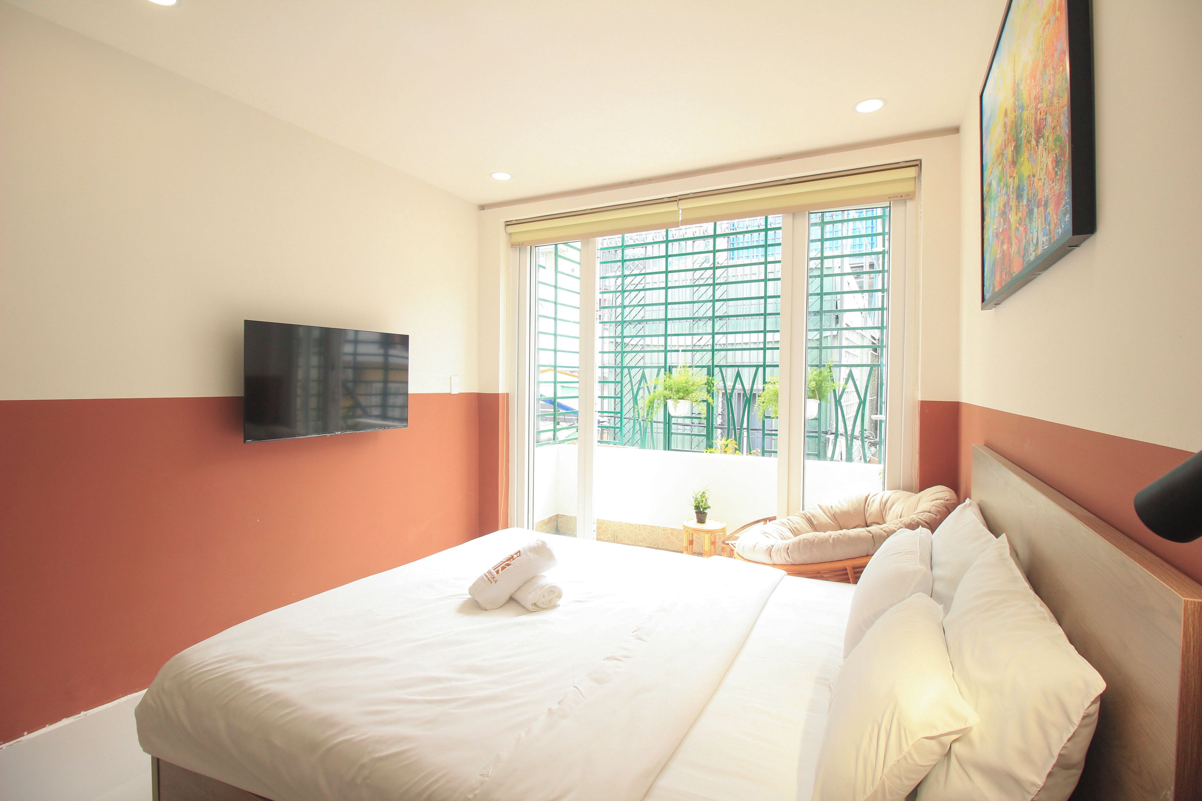 A hotel room with a bed, a television mounted on the wall, and a window with a view of buildings and greenery.
