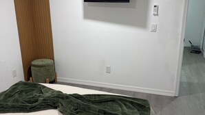 Quarto com cama, manta verde, televisão fixada na parede e painel de madeira à esquerda.