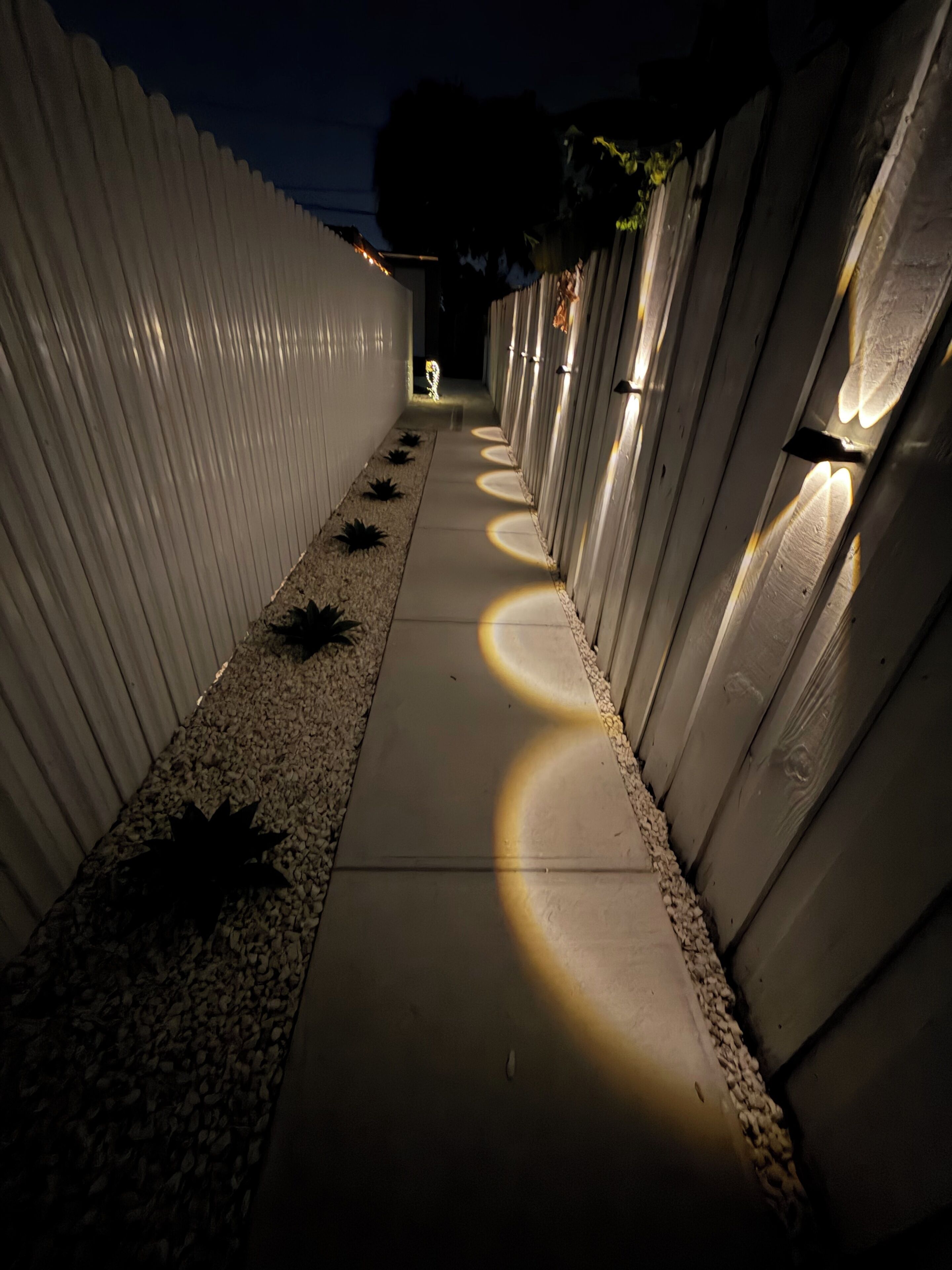 Ein Weg, der von weißen Mauern gesäumt wird, mit kreisrunden Leuchten, die Schatten auf den Boden werfen.
