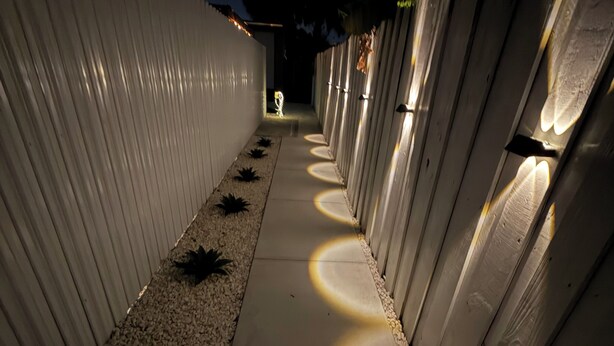 Um caminho ladeado por paredes brancas, com luminárias circulares que projetam sombras no chão.