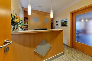 A reception area with a wooden reception desk, pendant lighting, a vase with flowers, and a door with a frosted glass panel. - Hotel Peterhof (Dietenheim)