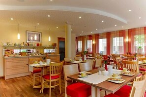 A spacious dining area with multiple tables and chairs, a kitchen area with a counter and appliances, and a television mounted on the wall. - Hotel Peterhof (Dietenheim)