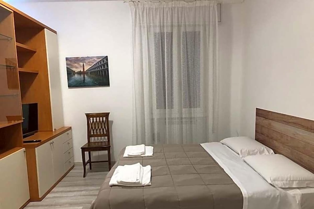 A hotel room with a bed, a chair, a TV, and a painting on the wall.