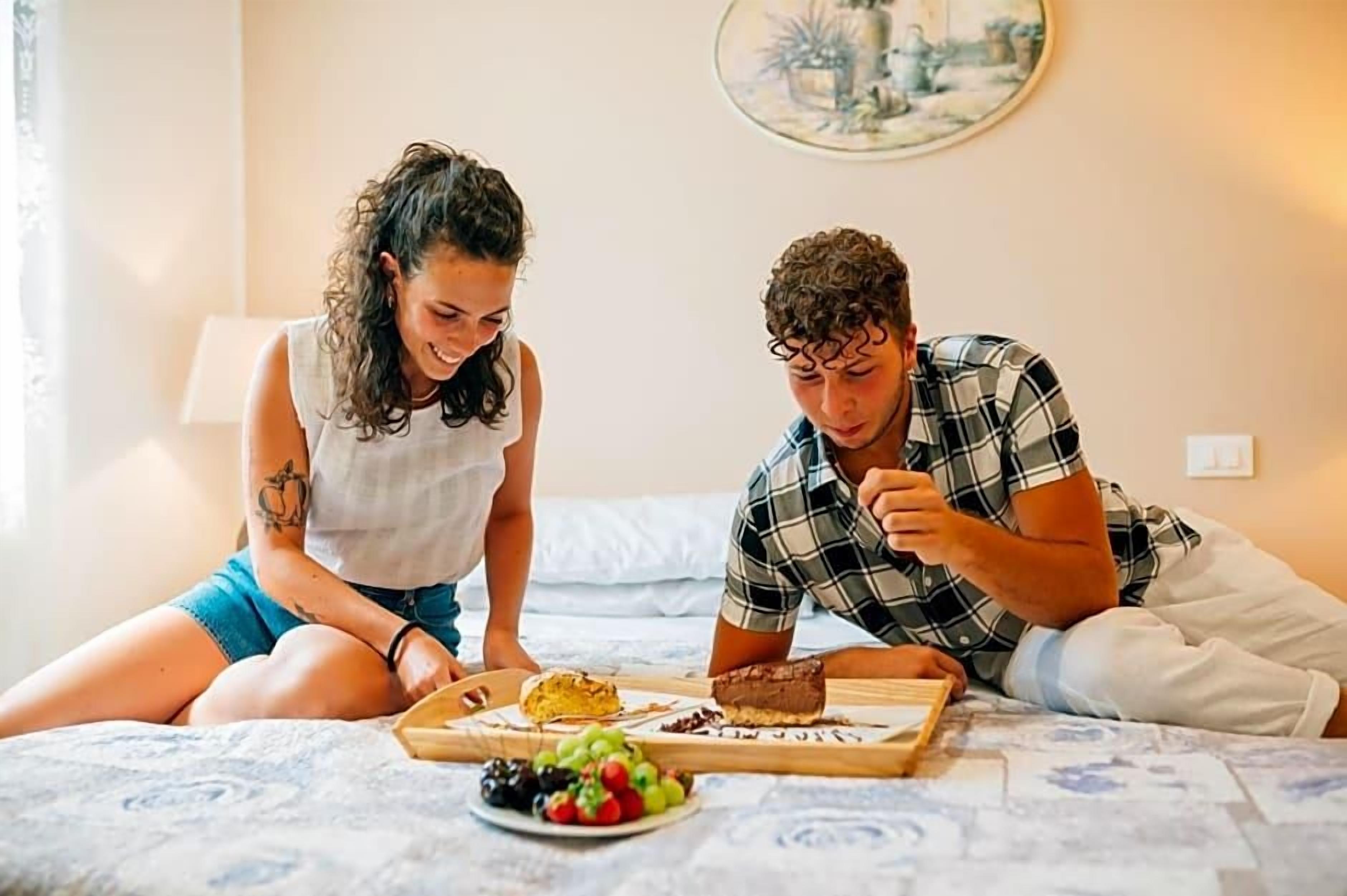 Zwei Personen sitzen auf einem Bett mit einem Frühstücksbrett, das Brot, Obst und eine Schokoladentafel beinhaltet.