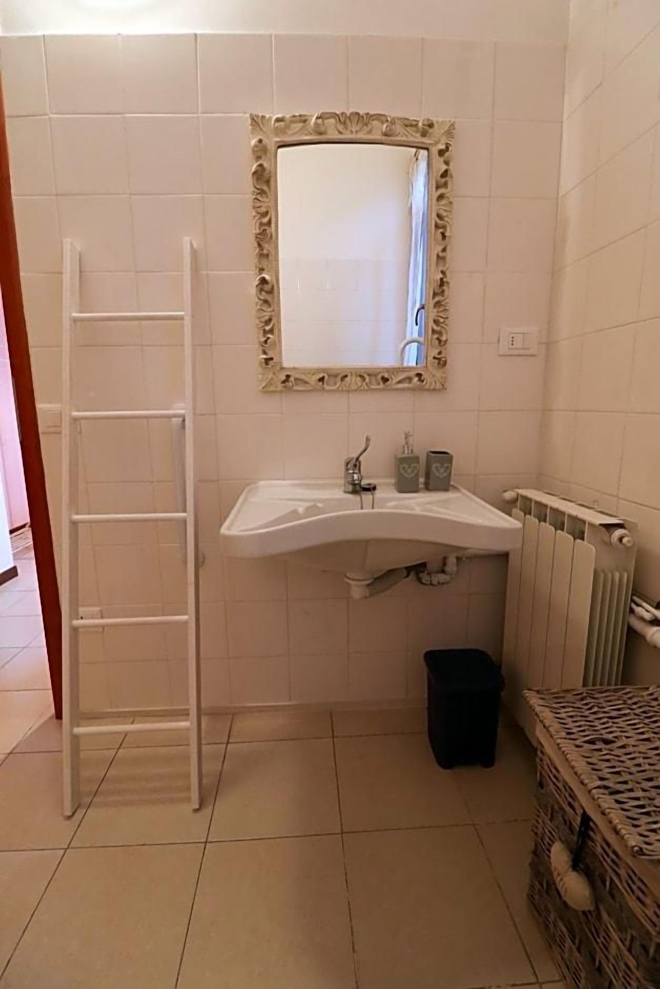 A bathroom with a white sink, a mirror with an ornate frame, a radiator, and a black trash bin.