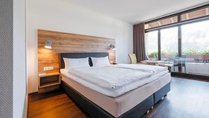 A modern hotel room with a large bed, a wooden headboard, a nightstand, and a view of the outdoors through large windows. - Restaurant Landhotel Winter (Gomadingen)