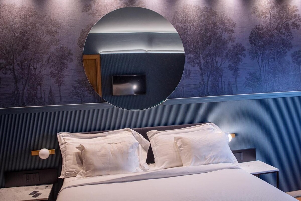 Habitación de hotel con cama, almohadas, mesita de noche, espejo y pared decorada con un diseño de árbol.