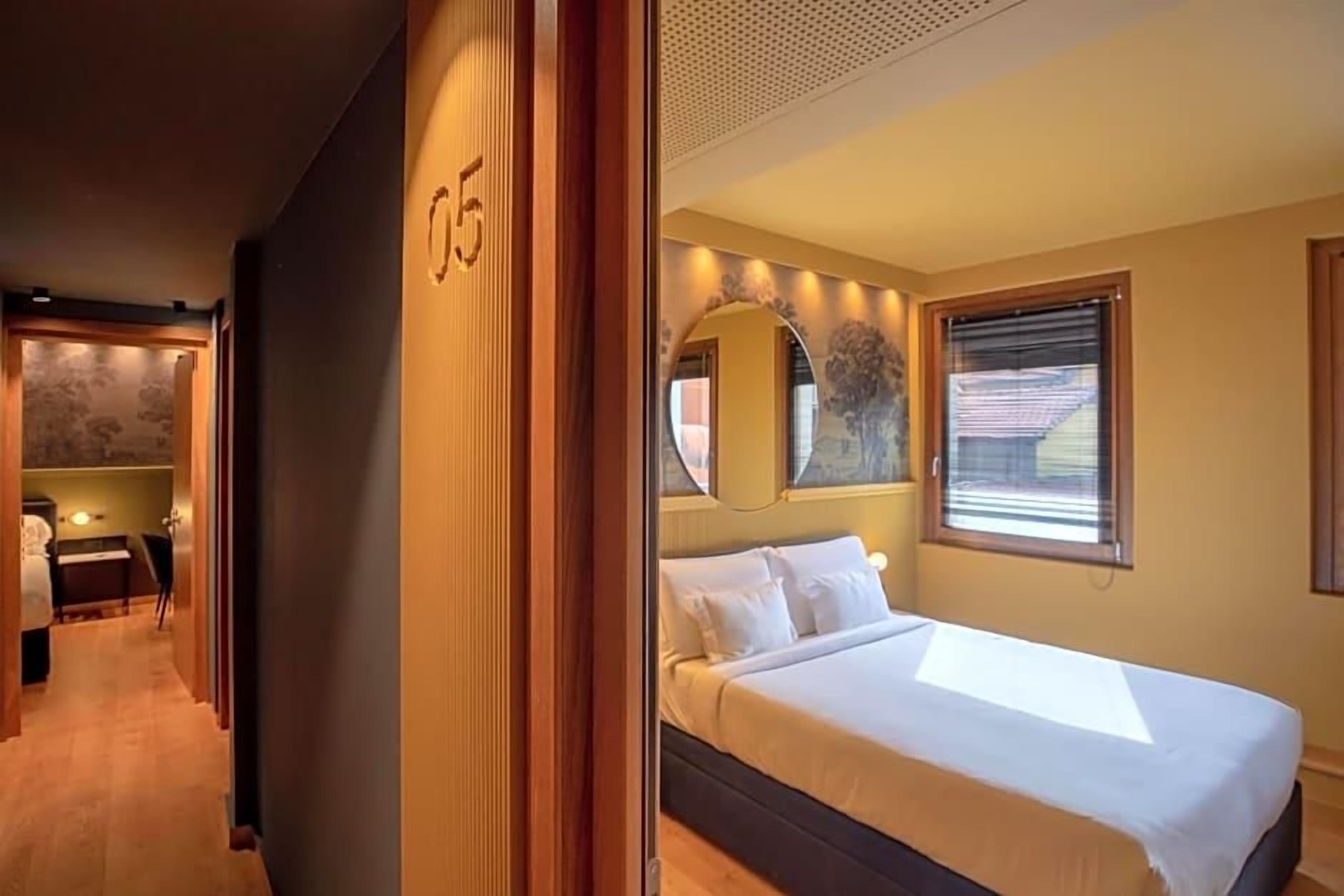 Una camera d'albergo con un letto, una finestra con persiane, uno specchio e una porta con un numero.