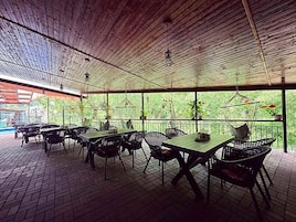 Un área de patio al aire libre con mesas y sillas, techo de madera y vista a follaje.