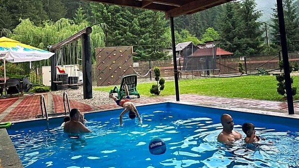 Piscina externa com área coberta, cercada por vegetação e um muro.