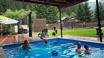 An outdoor swimming pool with a covered area, surrounded by greenery and a fence.