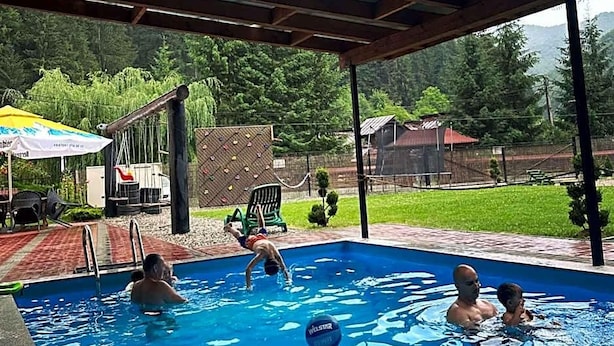 An outdoor swimming pool with a covered area, surrounded by greenery and a fence. - Maison Platanus (Voineasa)