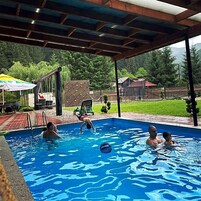 Piscina exterior con zona cubierta, rodeada de vegetación y una valla.