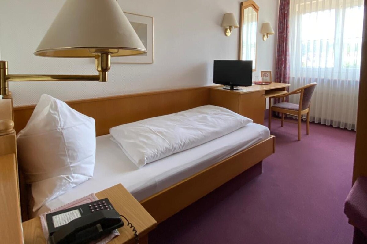 A hotel room with a single bed, a desk with a computer, a telephone, and a window with curtains.
