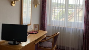 Quarto de hotel com escrivaninha, cadeira, televisão, duas luminárias de parede, um espelho grande e uma janela com cortinas finas.