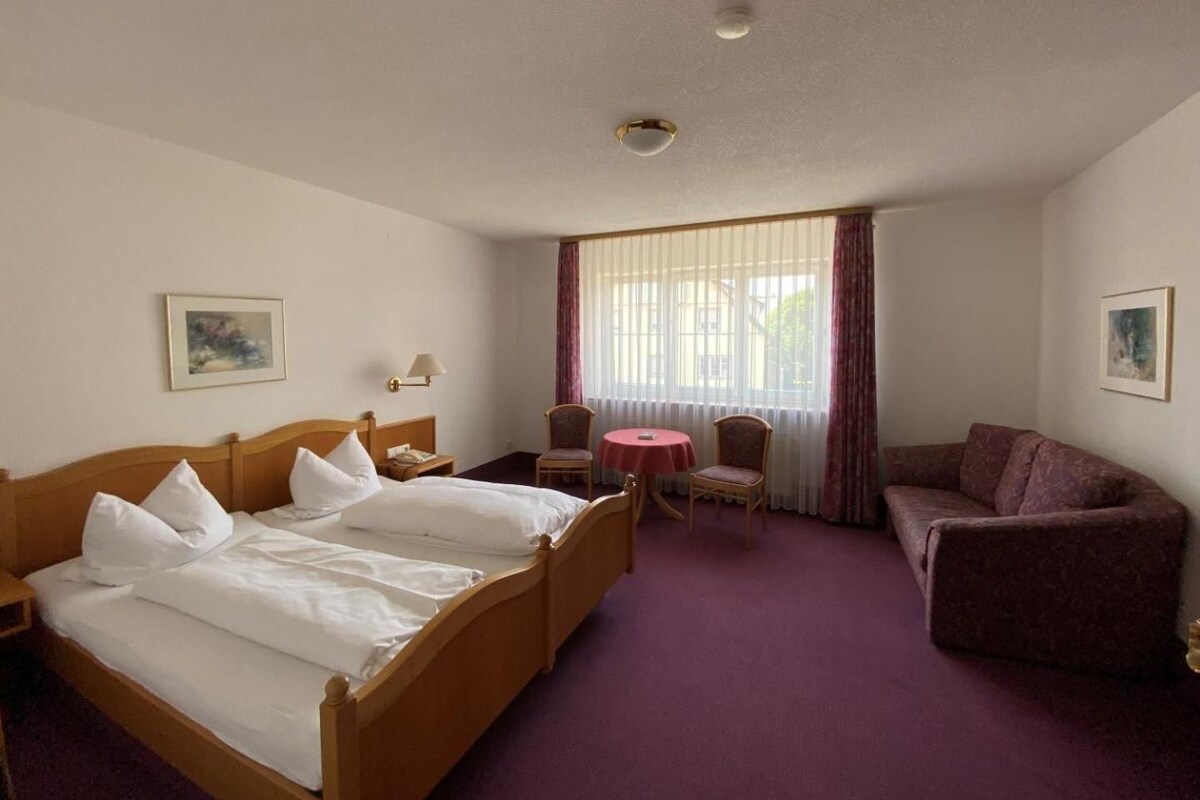 A hotel room with a bed, a desk, a chair, a sofa, and a window with curtains.