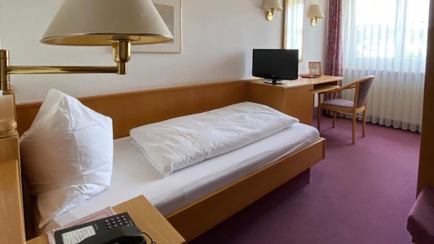 Quarto de hotel com uma cama de solteiro, uma mesa com computador, telefone e janela com cortinas.