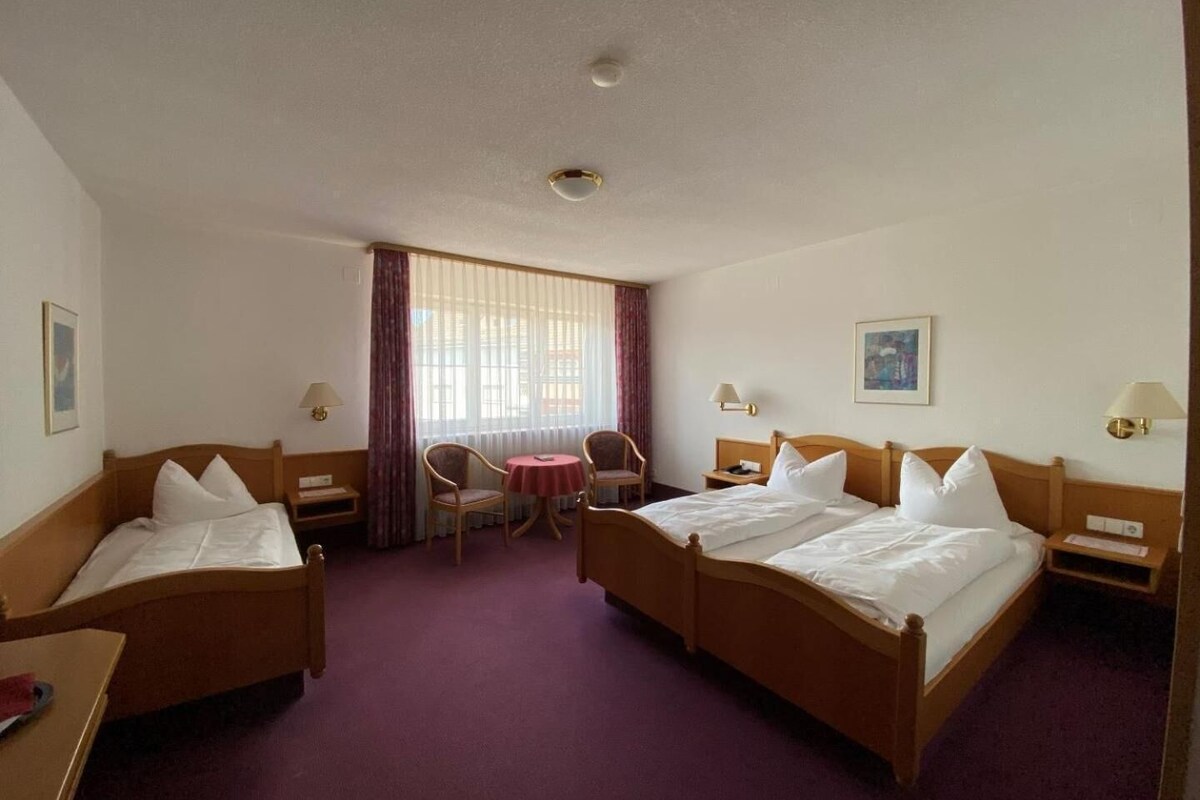 A hotel room with two beds, a small table, chairs, a window with curtains, and a painting on the wall.