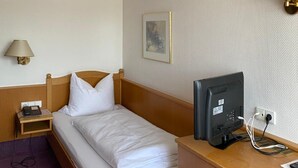 Quarto de hotel com uma cama de solteiro, mesa de cabeceira de madeira com abajur, escrivaninha com computador e luminária na parede.