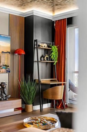 Una sala de estar moderna con una estantería en esquina, una lámpara de pie roja, una silla de madera y un cuadro de un paisaje urbano en la pared.