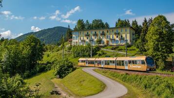 Un tren amarillo con 'Furnierstopp' en el costado, pasando por un hotel con balcones y un paisaje montañoso.