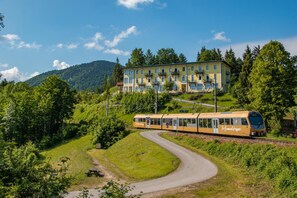 A yellow train with 'Furnierstopp' on the side, passing by a hotel with balconies and a mountainous backdrop.