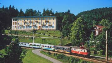 Ein Bahnhof mit einem Gebäude, das mehrere Fenster und Balkone aufweist, umgeben von Grünflächen und Hügeln.