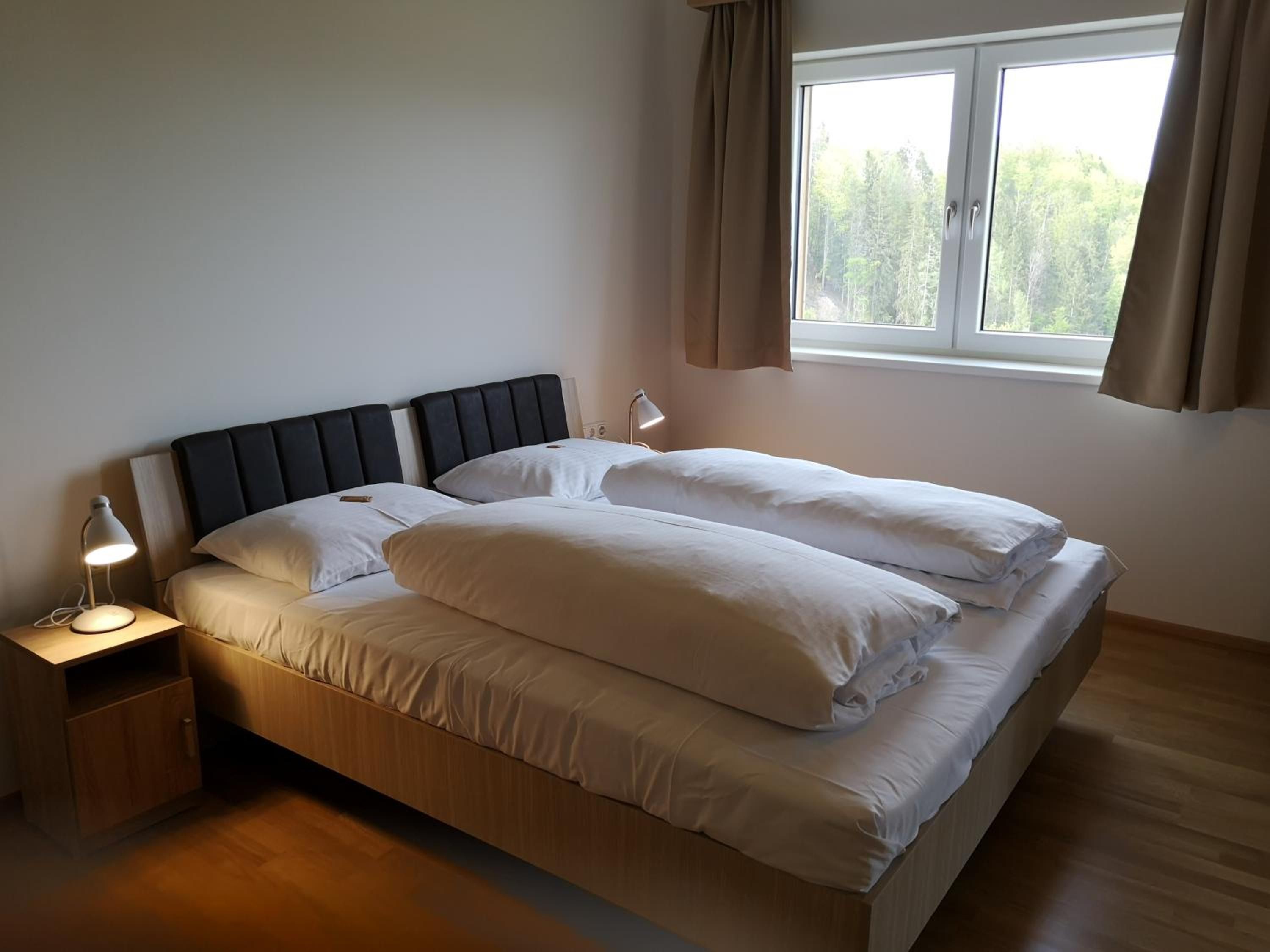 Ein Einzelbett mit Kopfteil, ein Nachttisch mit Lampe und ein Fenster mit Vorhängen.