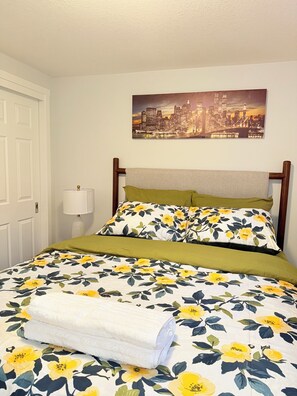 Una cama bien tendida con ropa de cama floral, un cuadro de un paisaje urbano encima y una lámpara al lado de la cama.