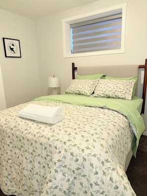 A neatly made bed with a floral bedspread, a pillow with a leaf pattern, and a folded white towel on top. - Brand new stylish basement suite. (Calgary)