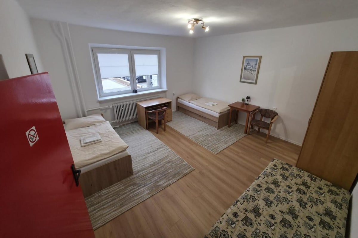 A small, sparsely furnished room with a bed, a desk, and a chair. There is a window providing natural light, and a radiator under the window.