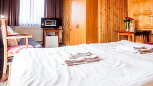Habitación de hotel con techo de madera, cama con sábanas blancas, televisor sobre un mueble de madera, una mesita con un florero y una ventana con cortinas.