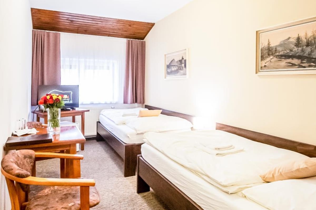 A hotel room with two single beds, a wooden desk, a chair, a small table with a vase of flowers, and a window with curtains.