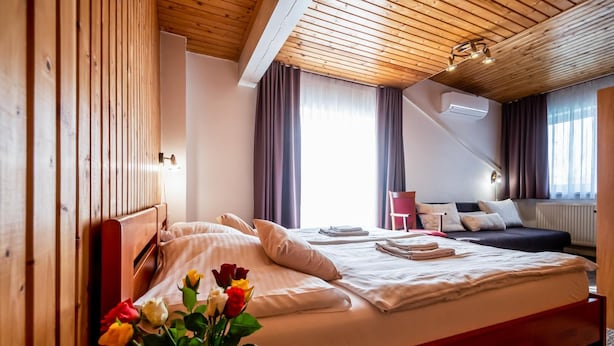 Habitación de hotel con techo de madera, cama con ropa blanca, cabecera roja, mesita de noche, escritorio, silla y ventana con cortinas.