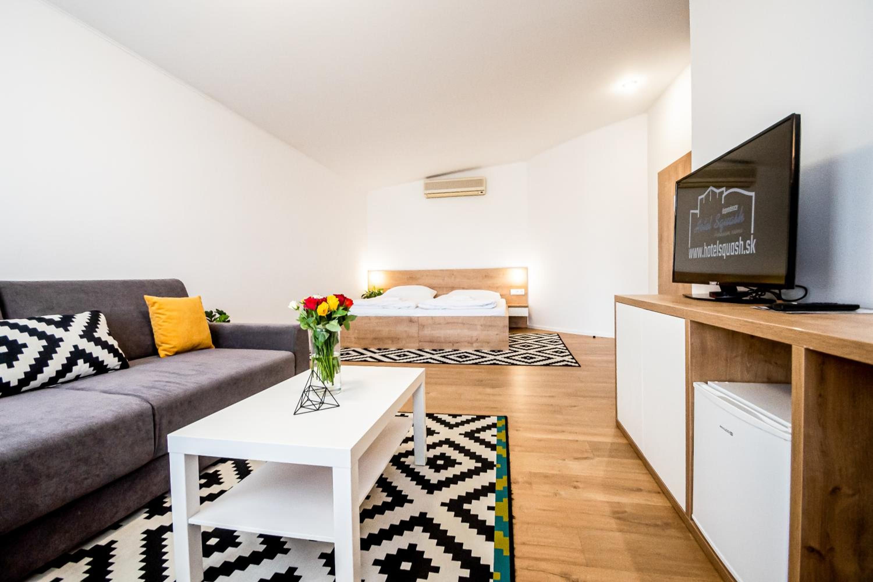 A modern hotel room with a grey sofa, a white coffee table, a bed with a wooden headboard, a TV on a wooden stand, and a small kitchenette.
