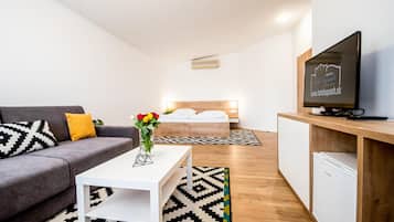 A modern hotel room with a grey sofa, a white coffee table, a bed with a wooden headboard, a TV on a wooden stand, and a small kitchenette.