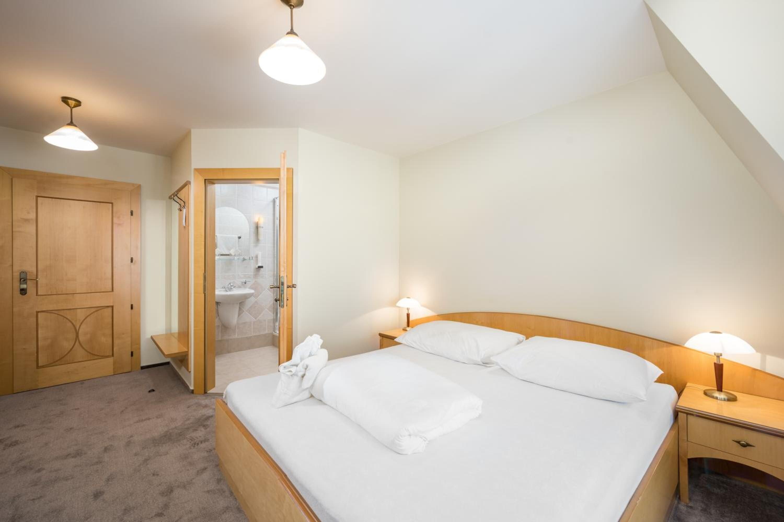 A bedroom with a bed, bedside lamps, a wooden door, and a bathroom visible through an open door.