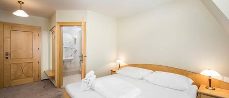 A bedroom with a bed, bedside lamps, a wooden door, and a bathroom visible through an open door.