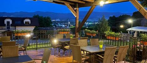 Un patio exterior con mesas y sillas, vistas de la ciudad y montañas al fondo.